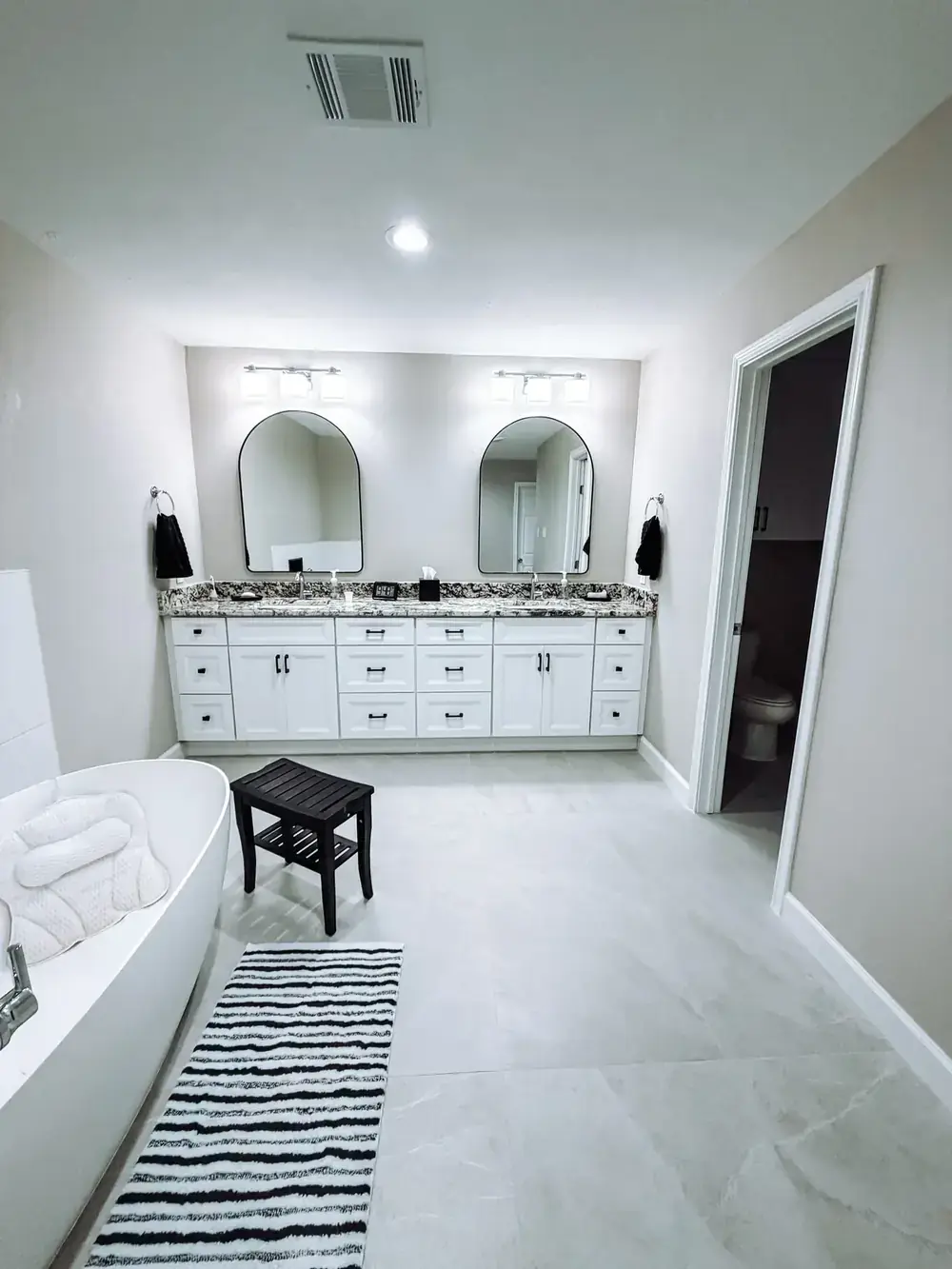 Master bathroom with a freestanding soaking tub, white double vanity with speckled granite, two arched mirrors, and a striped runner on large-format tile