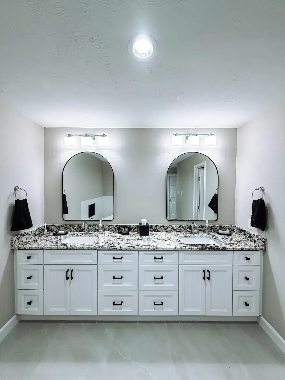 White shaker double vanity with speckled granite countertop, two arched black-framed mirrors, black sconces, and black hardware