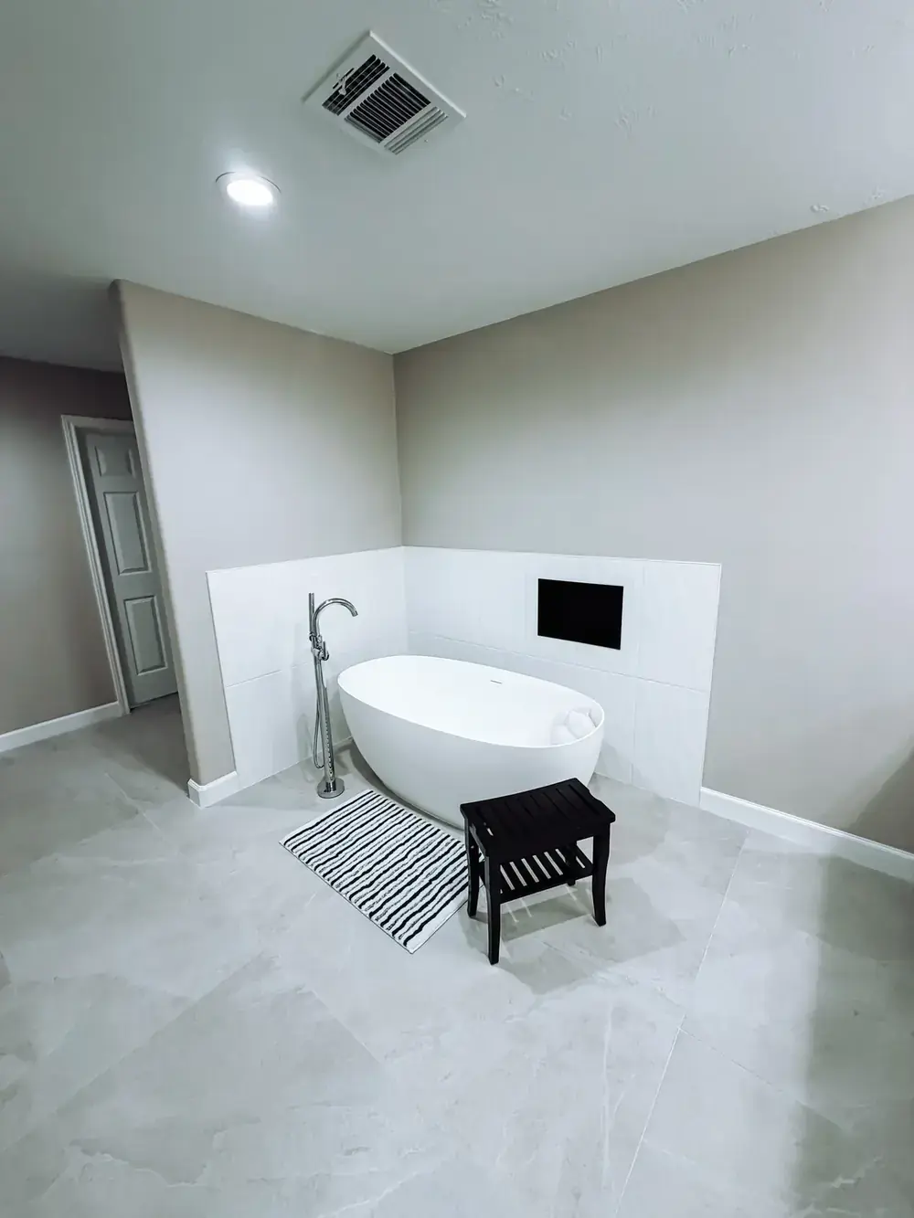 Freestanding tub set against a white tiled alcove with a recessed television, a dark teak stool, and large-format tile flooring