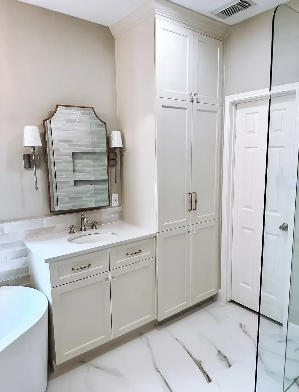 Bathroom with a greige vanity, arched mirror, wall sconce, floor-to-ceiling linen tower, freestanding tub, and marble-look tile floor