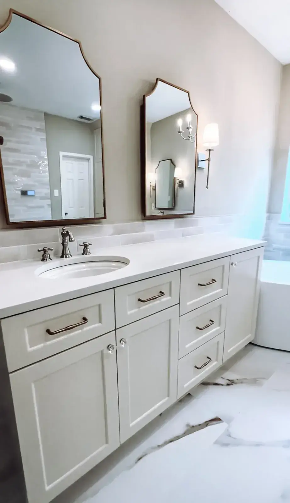 Refreshed bathroom with a white double vanity, brushed-gold fixtures, two ornate framed mirrors, and a tiled wall behind the tub