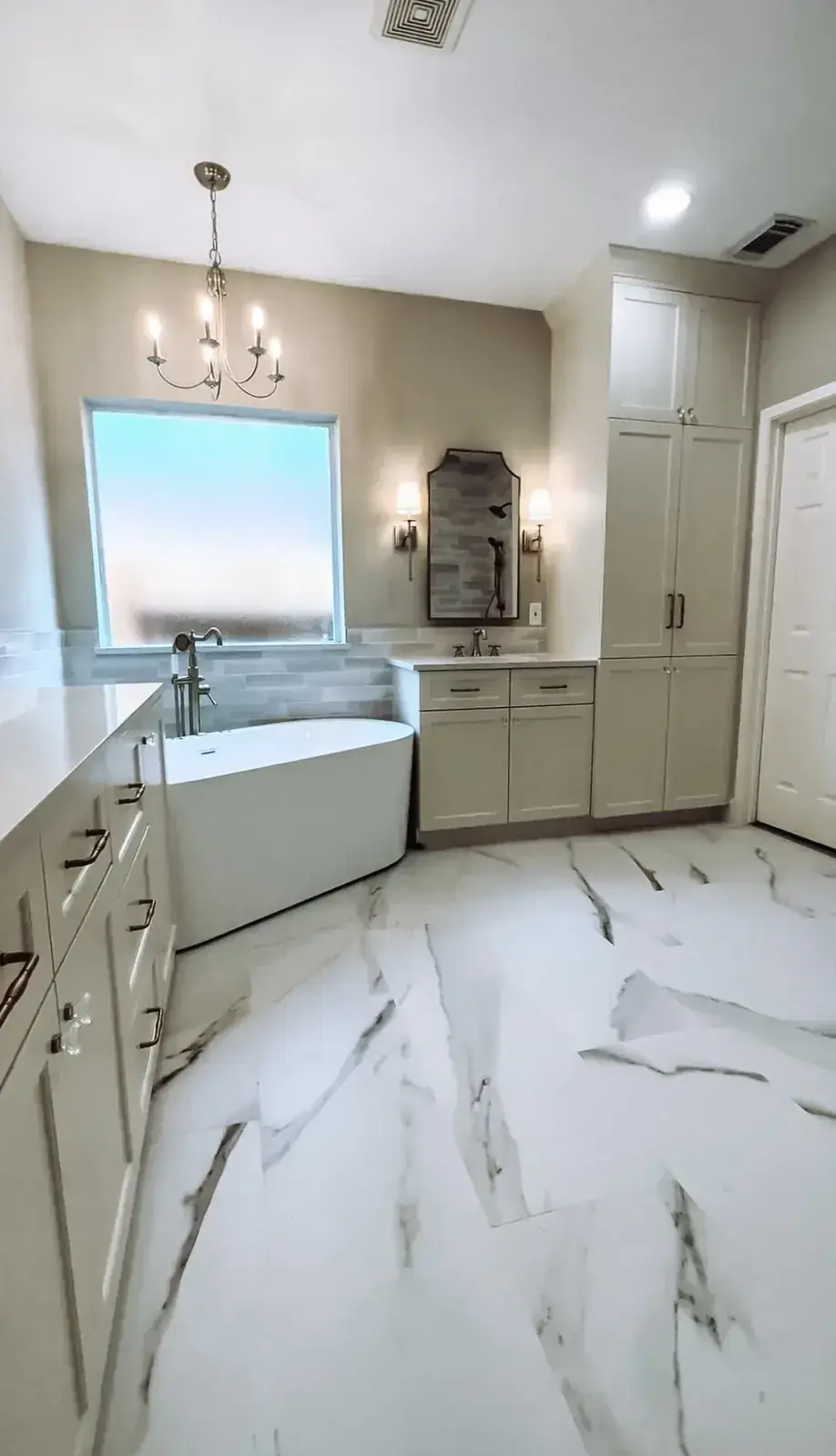 Freestanding tub beneath a frosted window with a candelabra chandelier overhead, sconces, and a large marble-look tile floor
