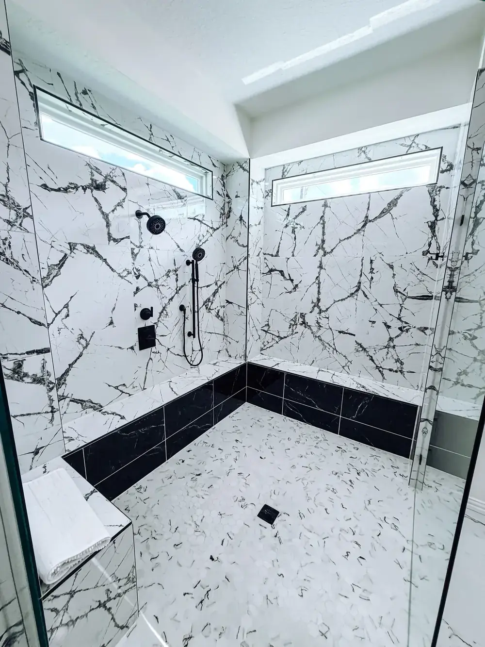 Walk-in shower with white marble-look slab walls, a black tile base and border, matte-black fixtures, and a high clerestory window