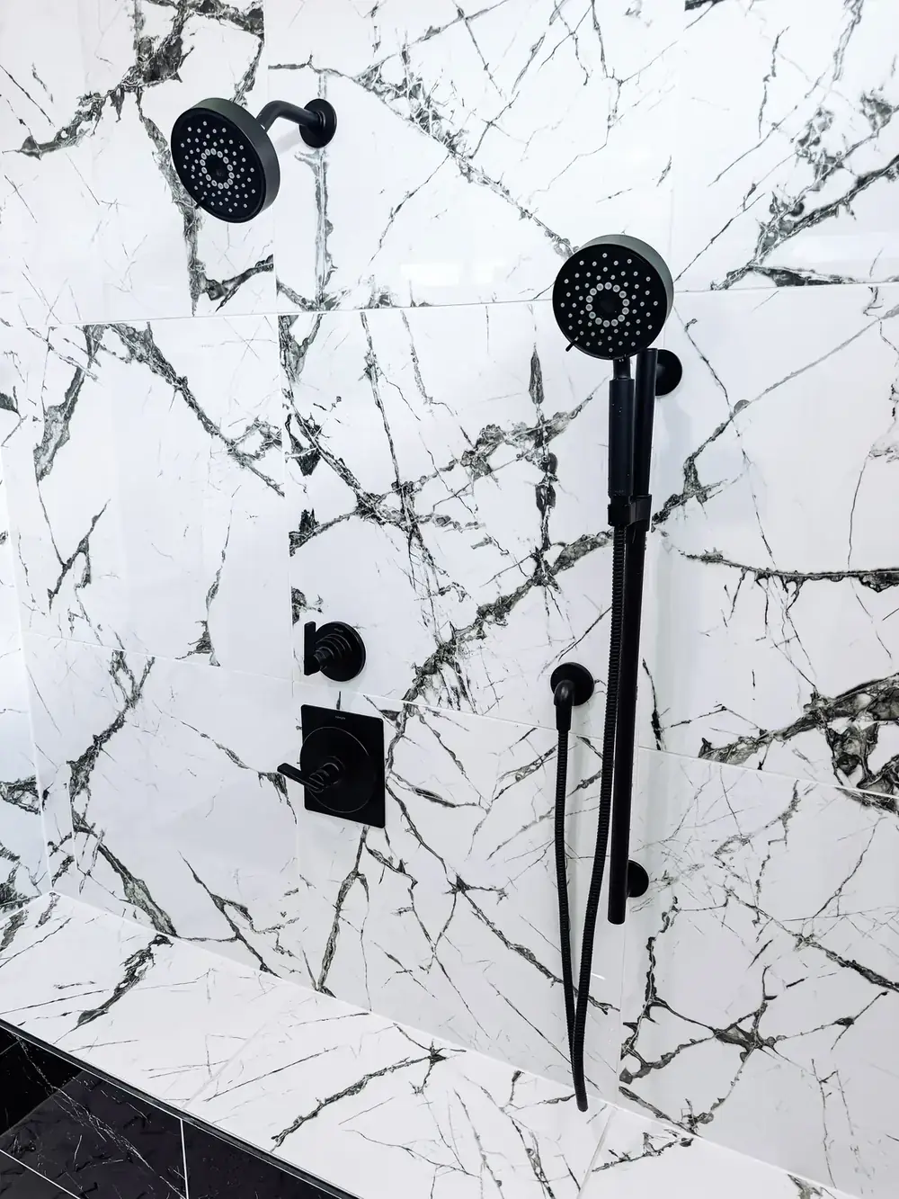 Close view of a marble-look shower wall with a matte-black rain head, handheld sprayer, and matching black controls