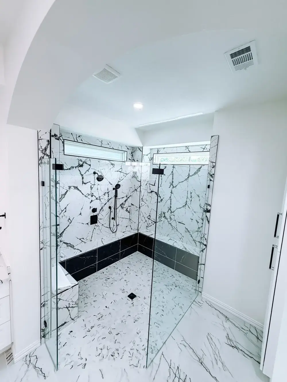 Large frameless glass walk-in shower with marble-look slab walls, black tile base, black fixtures, and clerestory windows above