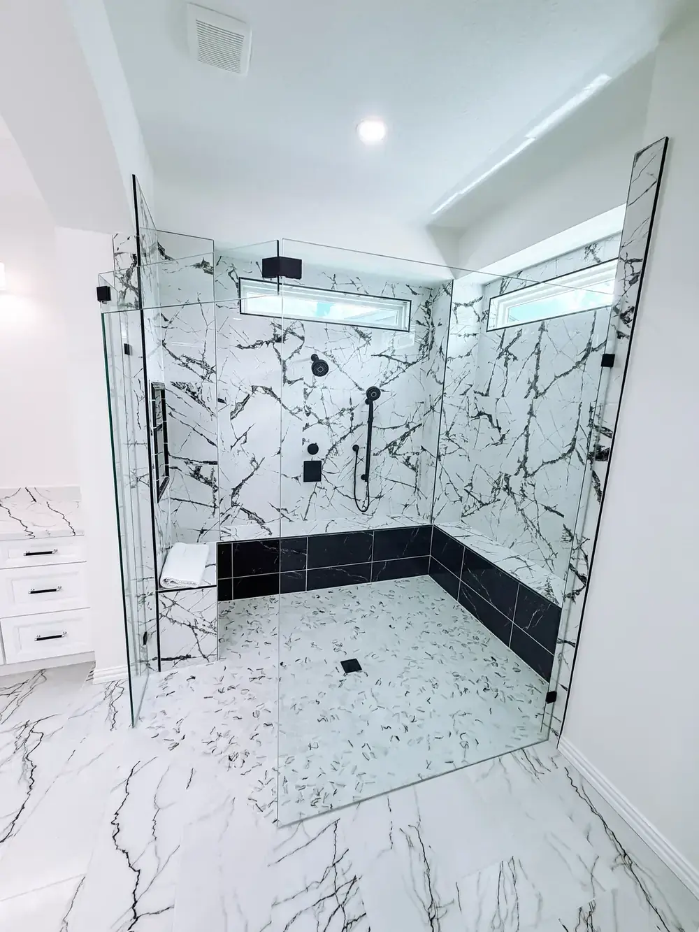 Large frameless glass walk-in shower with white marble-look slab walls, black tile shower base, matte-black fixtures, and clerestory windows