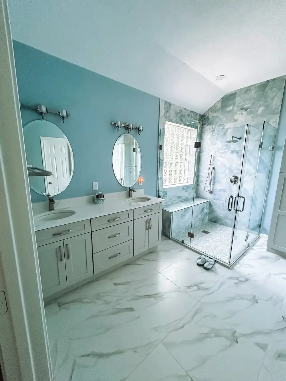 Bathroom with deep teal walls, a white double vanity with two round mirrors, a glass-block window, and a frameless glass shower with marble-look walls