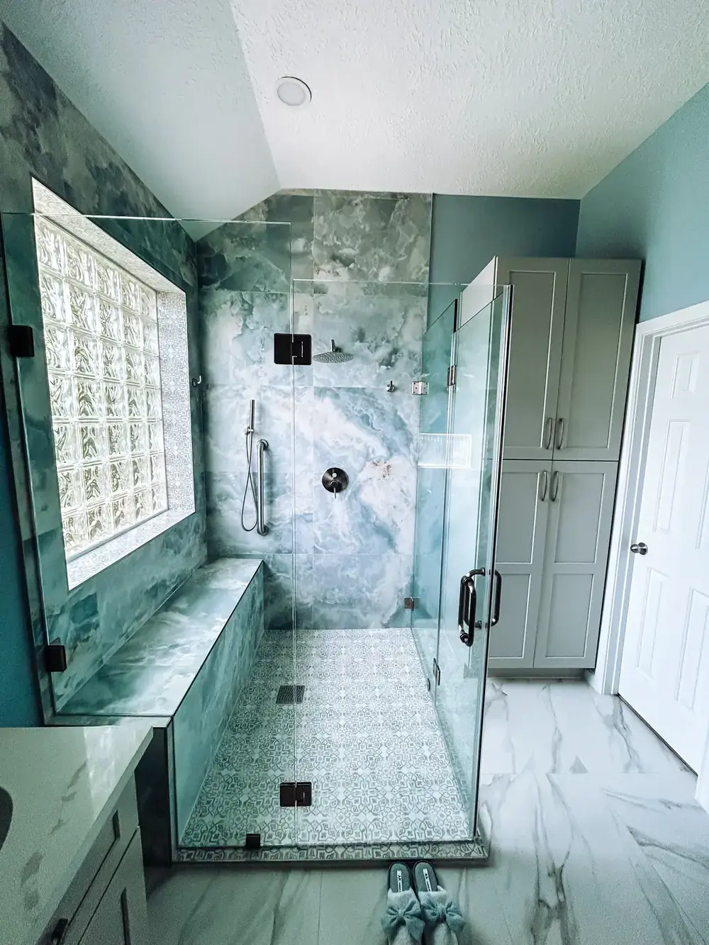Frameless glass shower with marble-look walls and a patterned mosaic shower floor, set against teal walls and a glass-block window