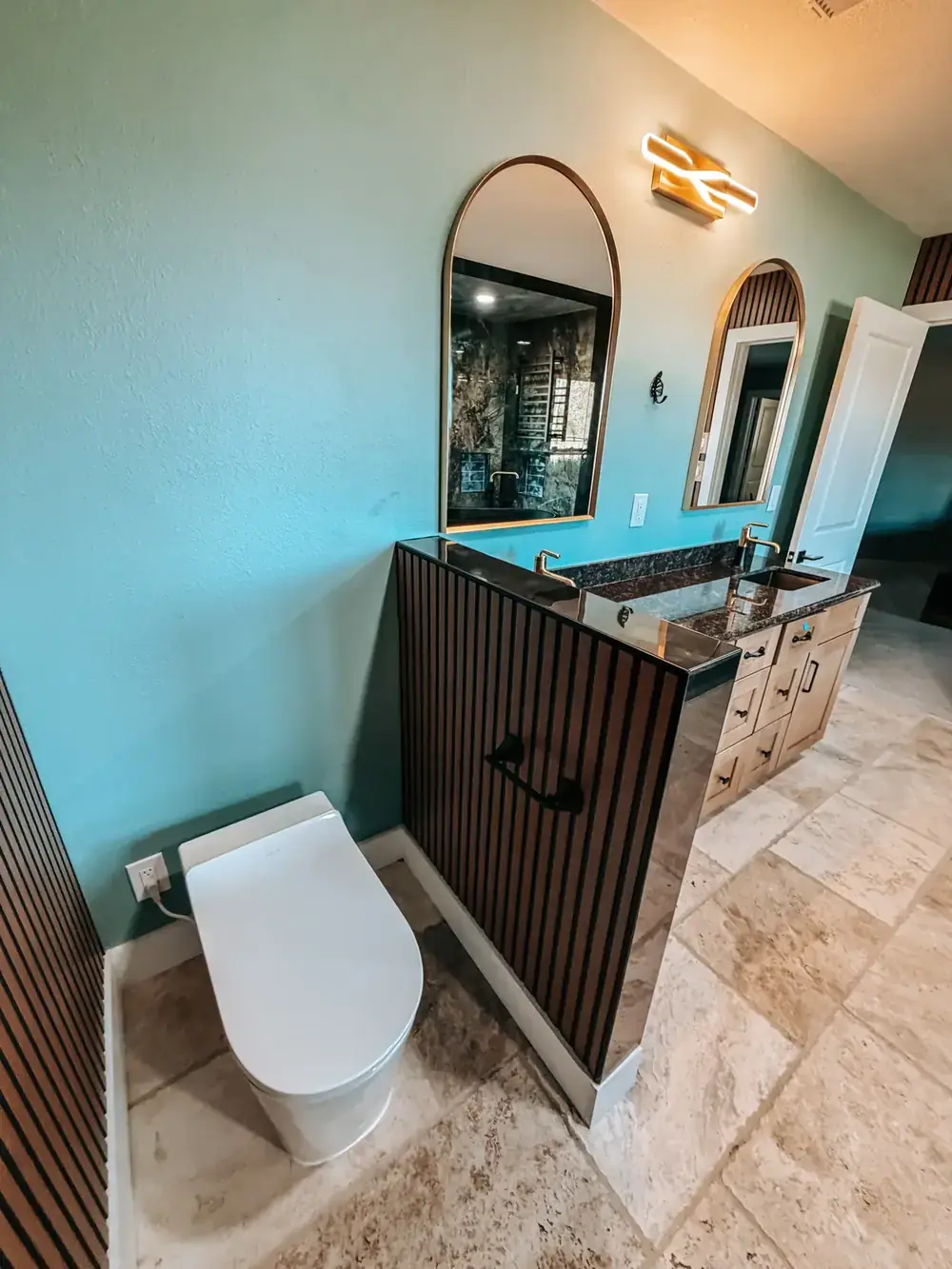 Remodeled bathroom with a deep teal accent wall, a fluted dark-wood vanity with a round mirror and sconce, and travertine-look tile flooring