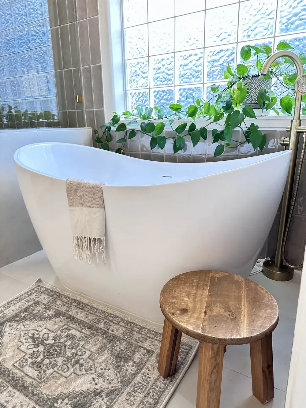 Freestanding slipper tub under a glass-block window with trailing plants, a patterned rug, and a round wood stool