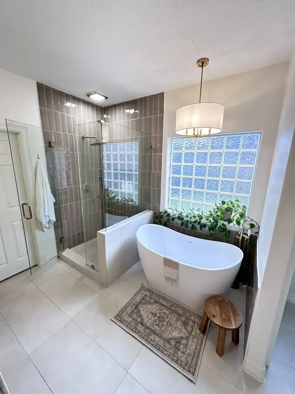 Bathroom with a grey tiled shower behind glass, a freestanding tub under a blue glass-block window, plants, and a drum pendant light