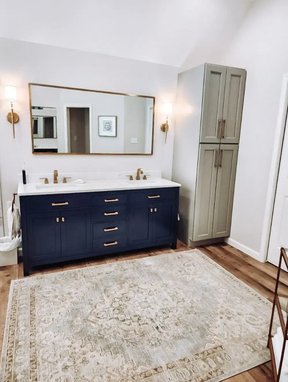Bathroom with a navy blue double vanity, brushed-gold hardware, a large gold-framed mirror, a tall greige linen cabinet, and a patterned rug