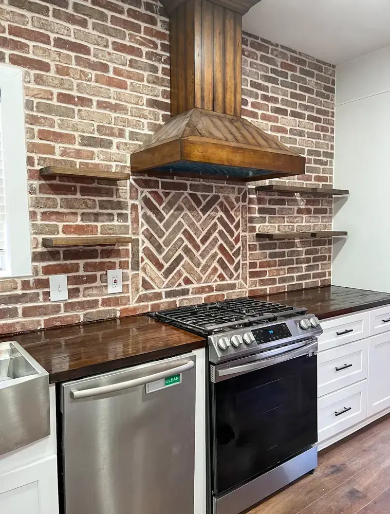 Exposed reclaimed-brick kitchen wall with herringbone inlay, custom wood range hood, and dark butcher-block countertops