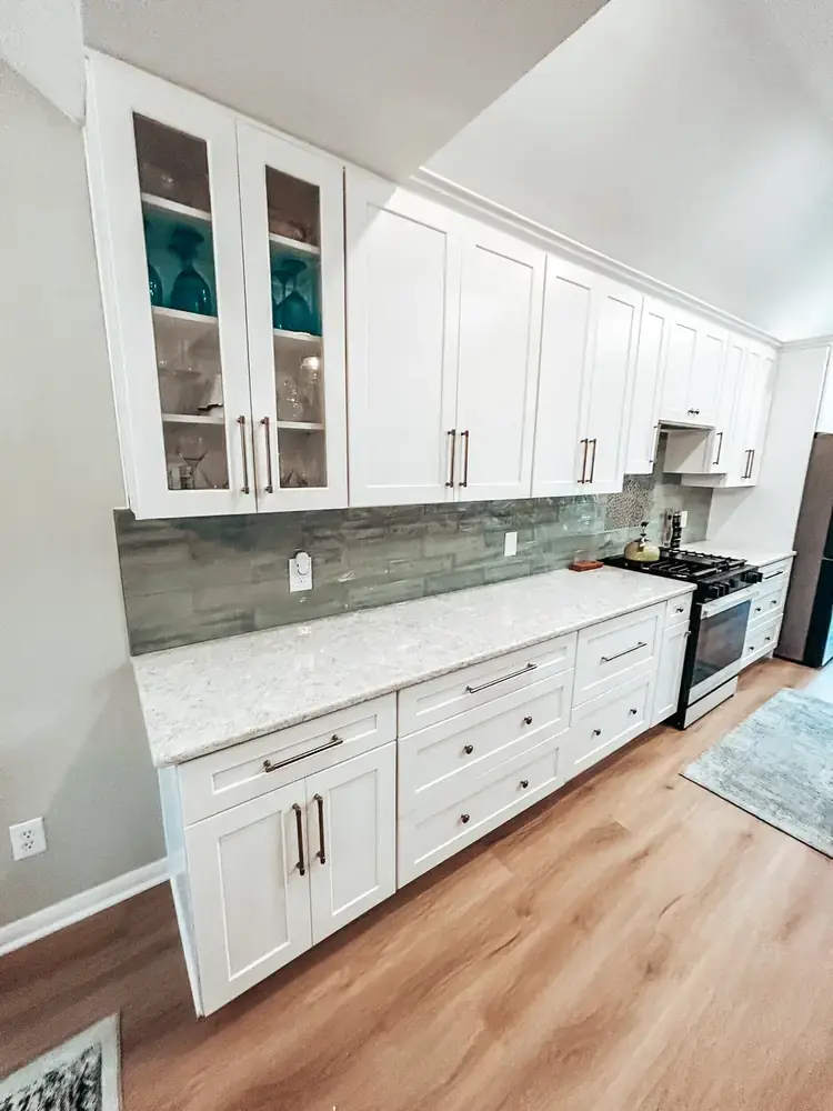 White kitchen with glass-front upper cabinets, sage-green glass tile backsplash, and pale quartz countertops